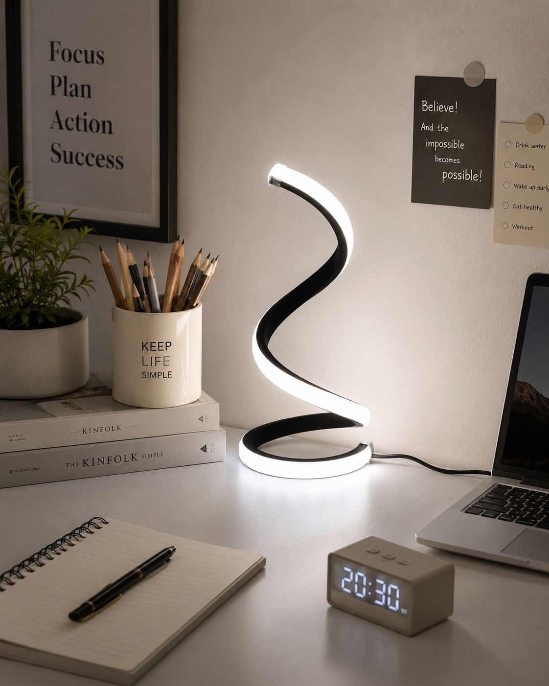 A modern spiral-shaped LED desk lamp with a black and white design, glowing on a white desk alongside a laptop, notebook, digital clock, and framed motivational posters.