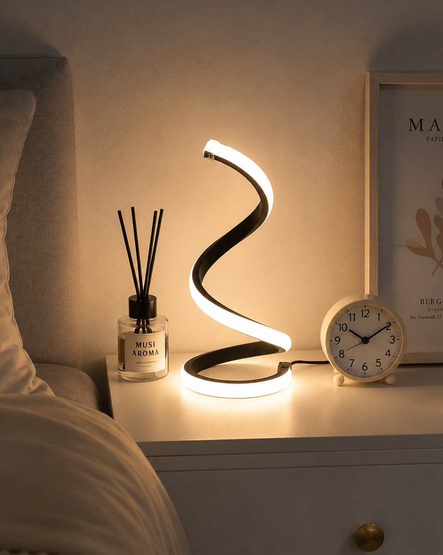 A modern spiral-shaped LED night light glowing with warm light on a white bedside table, styled in a bedroom next to a clock, diffuser, and framed art.